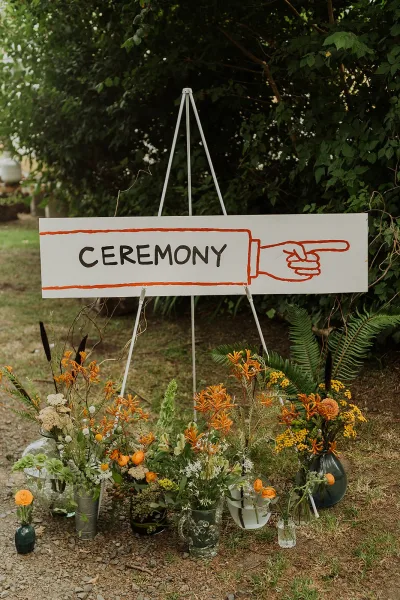 Wedding ceremony sign with painted lettering and pointing hand on an easel, surrounded by wildflower vases and ferns on a garden path