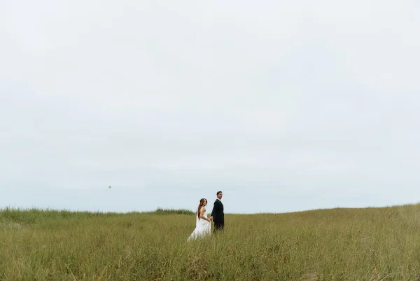 Couple portrait of bride and groom holding hands, her wedding dress and his suit in a tall grass field under an overcast sky