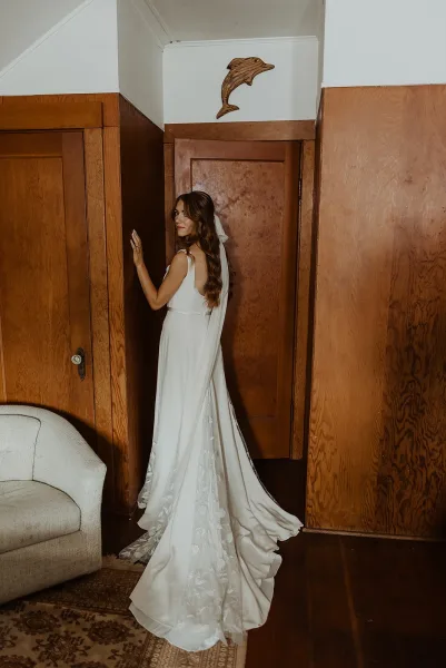 Bridal portrait of a bride looking back by a doorway, long veil trailing over a low-back wedding dress in a wood-paneled room