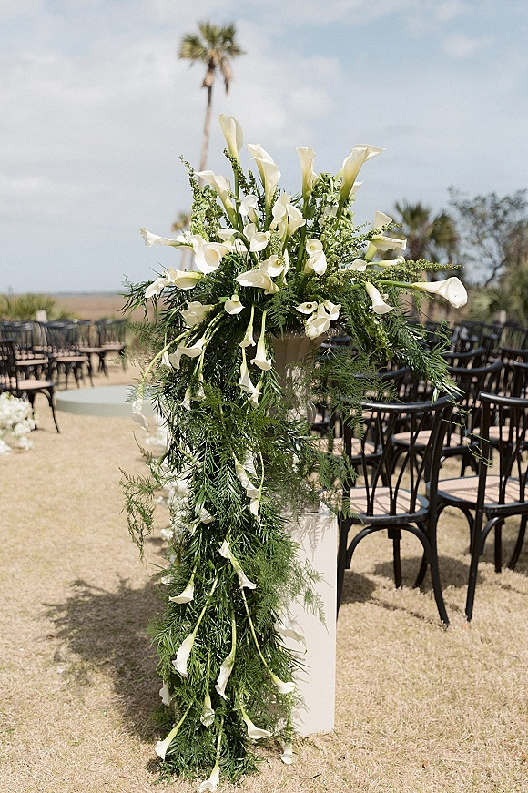 Ceremony floral arrangement of white calla lily ceremony flowers with greenery on a tall plinth lining an outdoor aisle with black chairs and palms