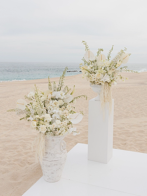 Ceremony floral arrangement of white anthurium, orchids, and roses with greenery on a tall pedestal by the ocean horizon on a sandy beach