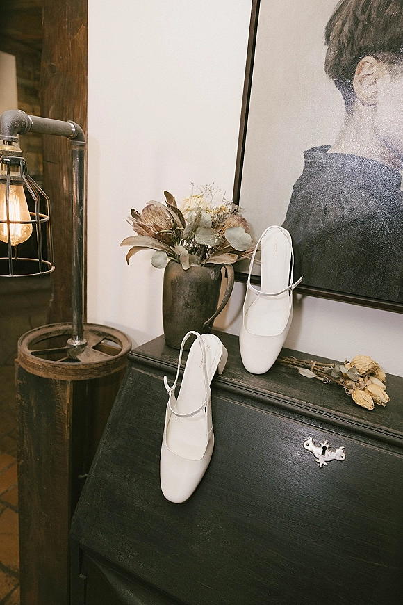 Wedding shoes, white slingback heels with dried flowers beside a ceramic vase and framed portrait on a wooden cabinet under an industrial lamp