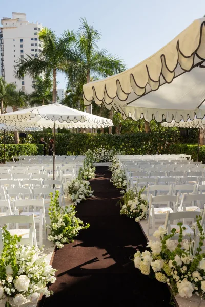 Ceremony aisle design with an outdoor ceremony aisle framed by white folding chairs, black runner, and white flower greenery arrangements under umbrellas