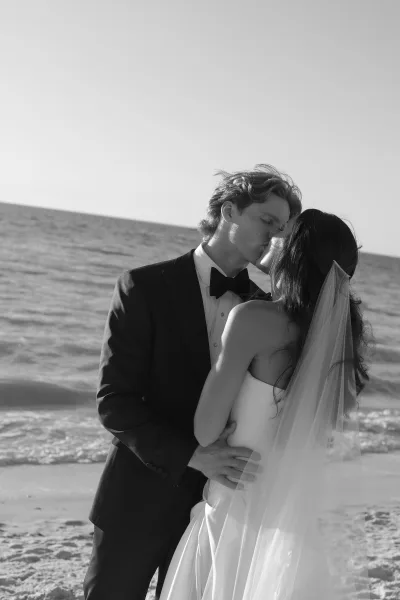 Wedding kiss in a black and white wedding photo, bride in strapless dress and veil kissing groom in tuxedo by the ocean shoreline