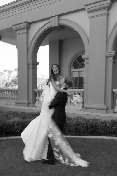 Couple portrait of groom carrying laughing bride in a strapless wedding dress and long veil, in front of arched estate columns