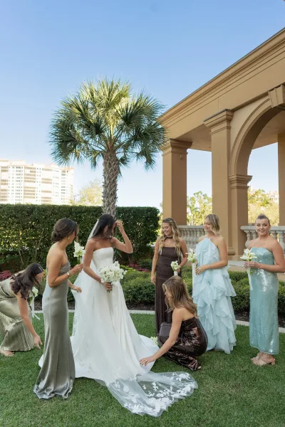 Bride with bridesmaids in a bridal party portrait as they adjust her lace veil train, holding white bouquet on a sunny lawn with palms