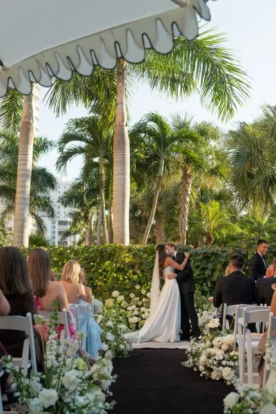 Ceremony kiss as bride in strapless gown and groom in tuxedo kiss under a hedge arch, with palm trees and blue sky behind