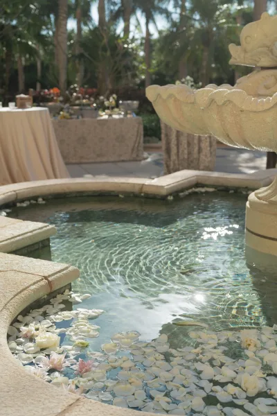 Wedding fountain decor with floating flower petals and roses in a stone basin, set in a palm-lined garden patio with cocktail tables
