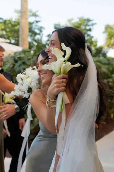 Bridal party moment as bridesmaids hug the bride, one holding a white calla lily bouquet with ribbon streamers in outdoor greenery