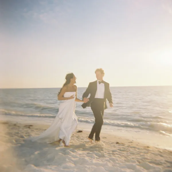 Couple portrait of newlyweds holding hands on a sunlit beach, bride in wedding dress and groom in tuxedo by ocean waves