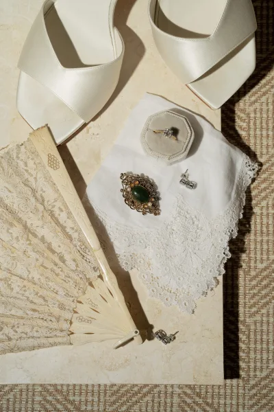 Bridal accessories flatlay with white satin heels and engagement ring box, lace fan and brooch arranged on stone and woven fabric