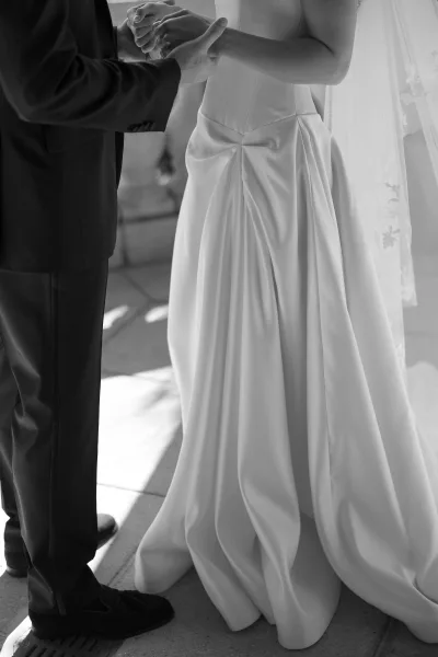 Wedding vows during ring exchange moment as groom places a band on bride’s hand, lace veil and tuxedo visible on stone floor backdrop