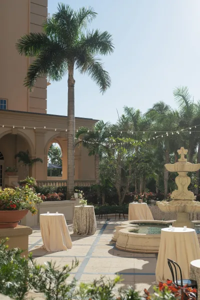 Outdoor cocktail hour with wedding cocktail tables in an arched courtyard patio, ivory linens and candles around a fountain under string lights