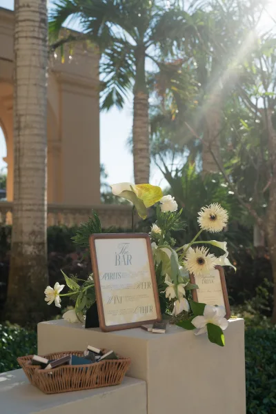 Wedding bar sign and wedding bar menu in wood frames on a pedestal, styled with calla lilies and greenery on a sunny patio with palm trees