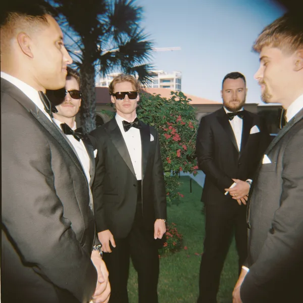 Groomsmen portrait of men in black tuxedos with bow ties and sunglasses, standing on a sunny lawn with palm trees and blue sky