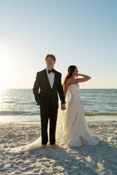 Couple portrait at the beach wedding portrait, bride in strapless gown and veil holding groom’s hand in black tux by sunlit waves