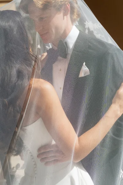 Wedding kiss portrait of bride and groom kissing under veil in warm indoor light, her strapless dress and his black tuxedo visible