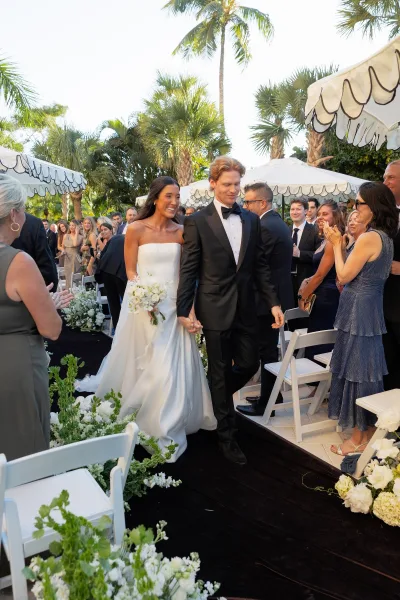 Wedding recessional as bride and groom walk the aisle hand in hand, bouquet raised, guests cheering on a palm-lined outdoor patio
