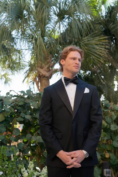 Groom portrait in a black tuxedo groom look with bow tie and pocket square, hands clasped in a garden with palm trees and sky