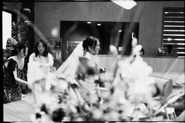 Wedding entrance as bride walks into indoor reception, smiling in veil with bouquet while bridesmaid carries her train past guests
