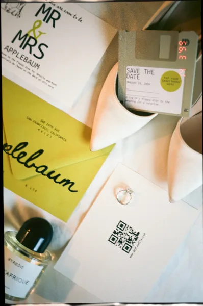 Wedding invitation suite with save the date card, green stationery, envelopes, QR code card, ring, perfume, heels, and floppy disk on white fabric backdrop