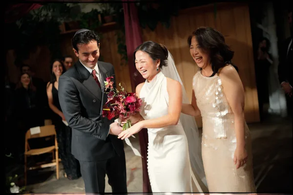 Ceremony moment as bride laughs holding a dark red bouquet beside groom in black suit, with veil and guests in an indoor space