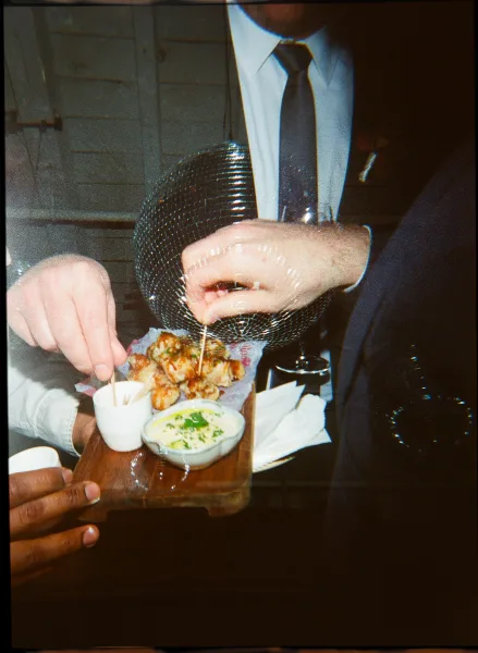 Reception appetizers on a wooden serving board with cocktail picks and dipping sauce, held by a guest in a dark suit by wood paneling