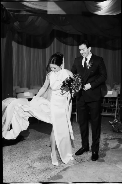 Ceremony recessional as newlyweds walking down aisle, bride in sleeveless gown and long veil holding bouquet under draped fabric ceiling
