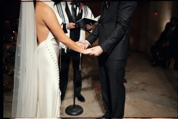 Wedding vows as bride and groom hold hands, her long veil and buttoned strapless dress beside his suit, tallit and mic in dim aisle ceremony