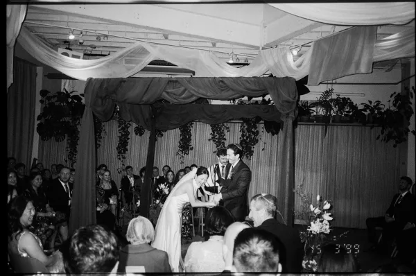 Ceremony moment as bride and groom stand under a chuppah with draped fabric and greenery, guests seated in an indoor venue with beams