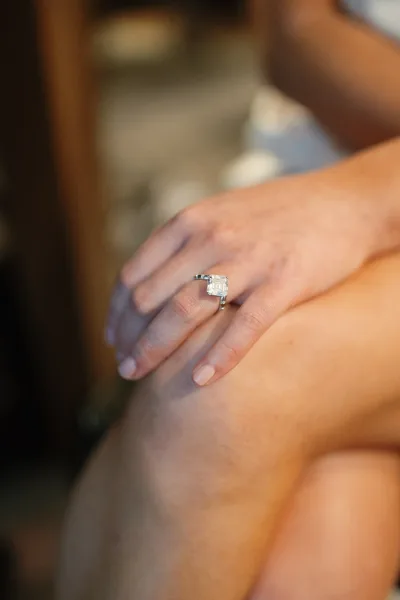 Engagement ring with an emerald cut diamond in a solitaire setting on manicured nude nails, against a warm blurred indoor background