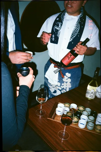 Sake tasting setup with a wedding sake toast, ceramic cups on a wooden tray beside a labeled bottle and wine glasses on a table