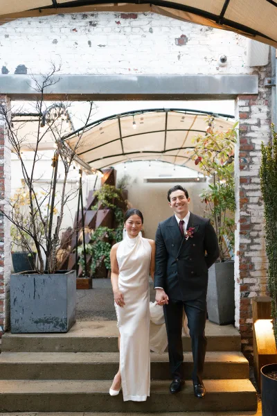 Couple portrait of bride and groom holding hands on concrete steps, her veil and slit dress beside his double breasted suit by a brick courtyard wall