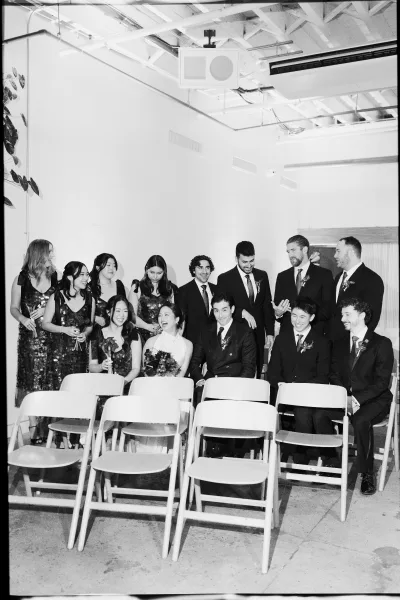Wedding party photo of bridesmaids and groomsmen seated on folding chairs, laughing with bouquets and boutonnieres against a white wall indoors