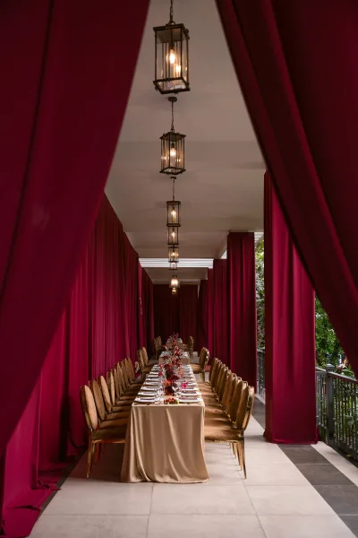 Reception tablescape with long banquet table wedding setup, burgundy draping overhead, candlelit florals and lantern lights on a covered terrace