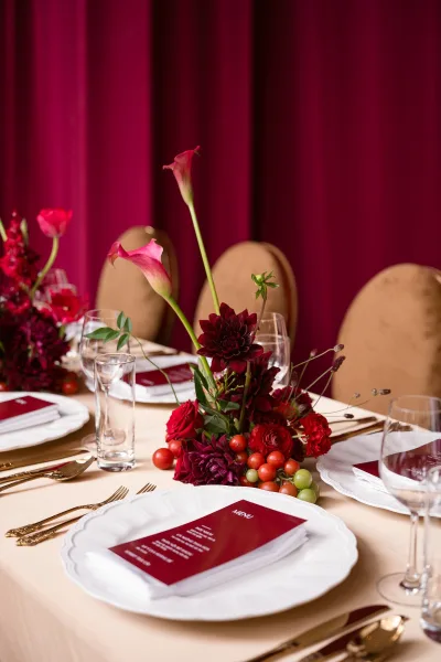 Reception tablescape with burgundy wedding tablescape florals—calla lilies, roses, dahlias, berries—gold flatware, menu card, burgundy drape backdrop