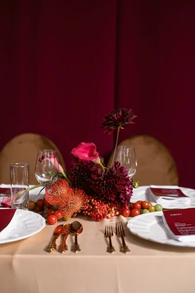 Reception tablescape with wedding table centerpiece of dahlias and calla lily, menus and glassware on neutral linens before a burgundy drape backdrop