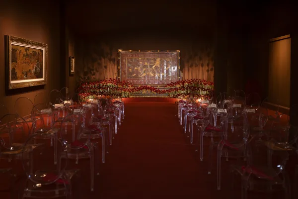 Ceremony setup for an indoor wedding ceremony with acrylic ghost chairs and a red floral aisle arrangement in a moody art gallery room