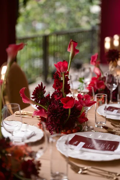Reception tablescape with a red wedding centerpiece of calla lilies, dahlias and grapes, gold flatware, and menus on an outdoor patio with greenery