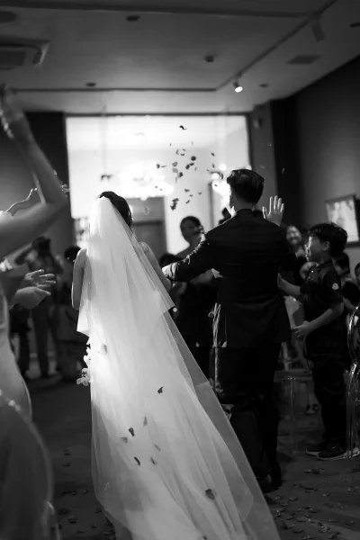 Wedding recessional as bride and groom walk out down the indoor aisle, bridal veil trailing while guests cheer and toss flower petals overhead