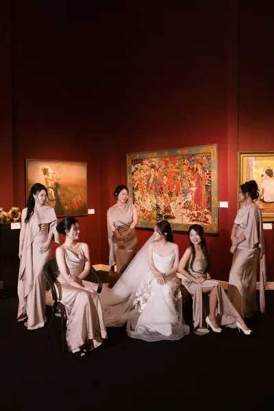 Bride with bridesmaids in satin champagne dresses, seated with bouquet and veil against a striking red wall in an art gallery interior
