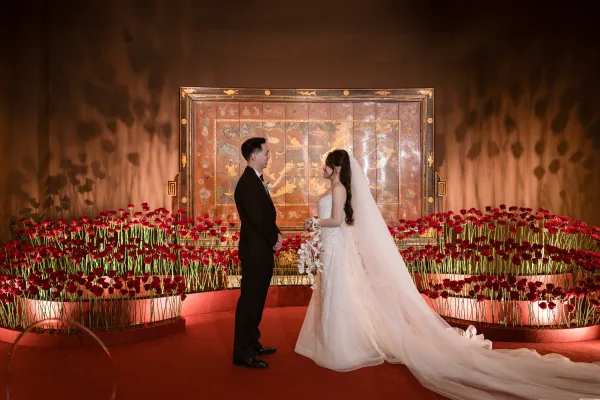 Couple portrait of bride and groom facing each other on an indoor stage, her veil and orchid bouquet framed by red rose backdrop