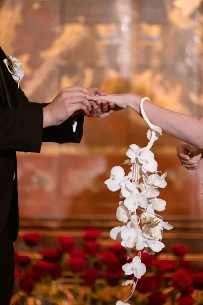 Ring exchange close up as groom in tuxedo places wedding band on bride’s hand, white orchid wrist corsage against warm indoor backdrop