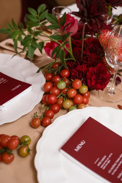 Reception tablescape with a red floral centerpiece, greenery, cherry tomatoes, wine glasses, and white scalloped plates on tan linen tablecloth