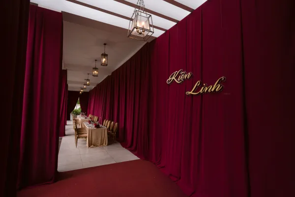 Reception tablescape with long banquet reception table, burgundy draping, lantern lights, beige linens, florals, and a gold script sign in a corridor
