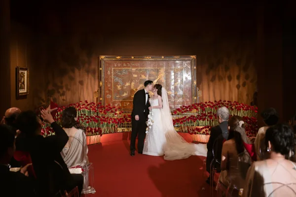 Ceremony kiss as bride in lace dress and long veil kisses groom in black tux beside red rose aisle florals in indoor room