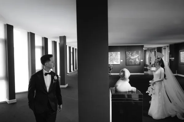 First look moment as groom in tuxedo walks toward bride in veil holding a white orchid bouquet in a gallery hallway with columns