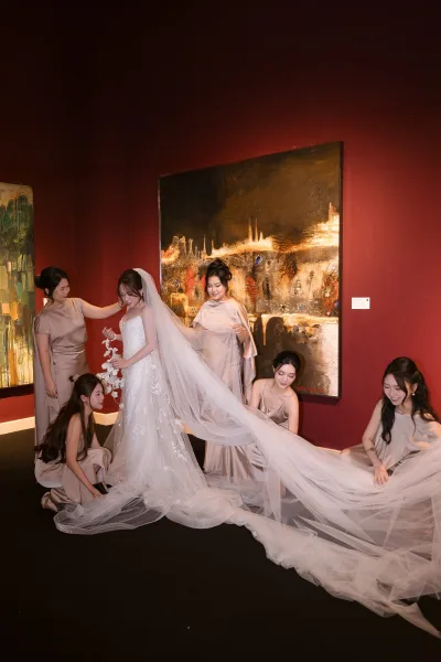 Bride with bridesmaids as they fix her long veil and strapless lace gown, standing before a red wall with framed abstract art in a gallery