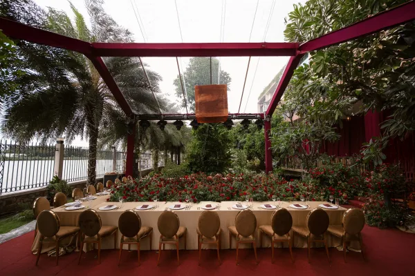 Reception tablescape with a long banquet table wedding setup, red floral runner and gold chairs under hanging lanterns by the riverside garden