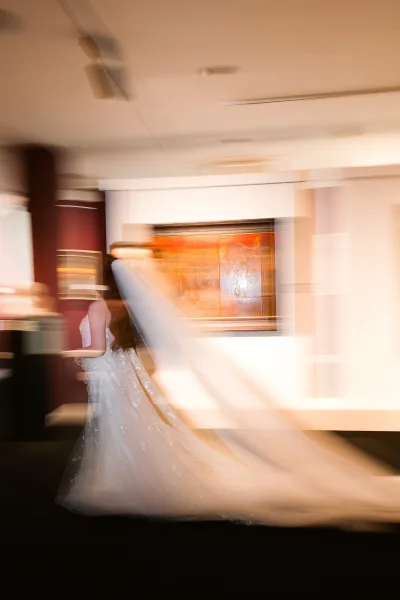 Bridal portrait of a bride with long veil in a strapless lace gown, looking back while walking through an indoor hallway with wall art